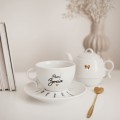 Biały zestaw porcelanowy z personalizacją - Imbryk i filiżanka ze spodkiem z imionami na zakończenie roku Zbliżenie na złote zdobienie na białym filiżance porcelanowej imbryk i filiżanka z imionami dzieci na spodku nadzwyczajnie.mom